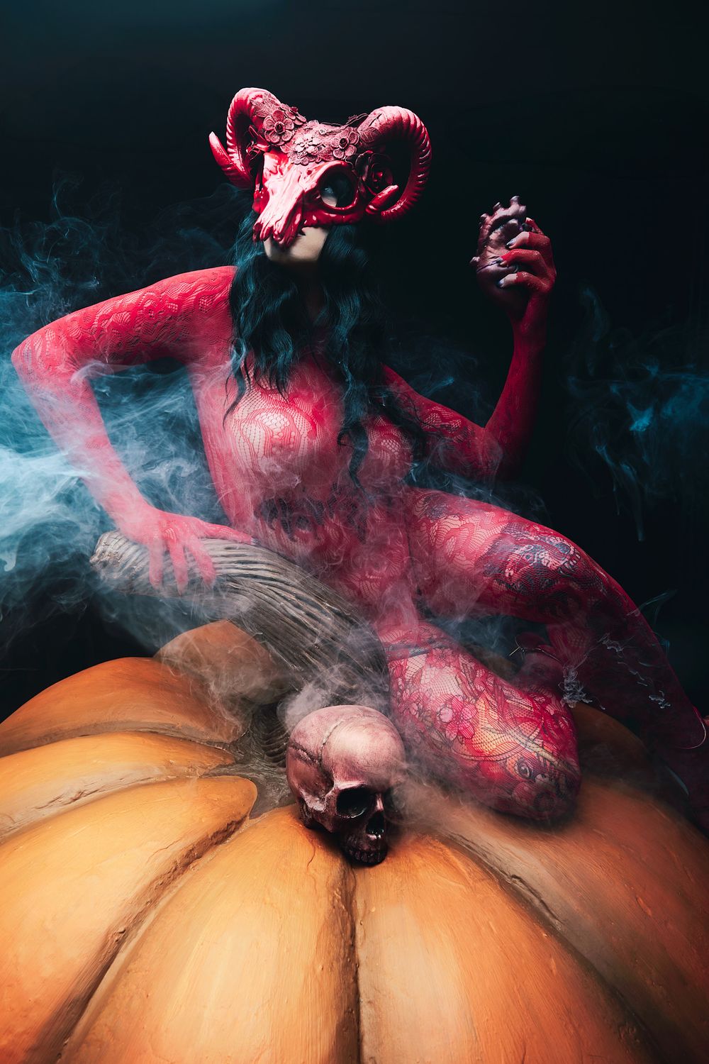 Red demon figure wearing ram skull mask sits atop pumpkins with skull, surrounded by mystical smoke in Halloween art.