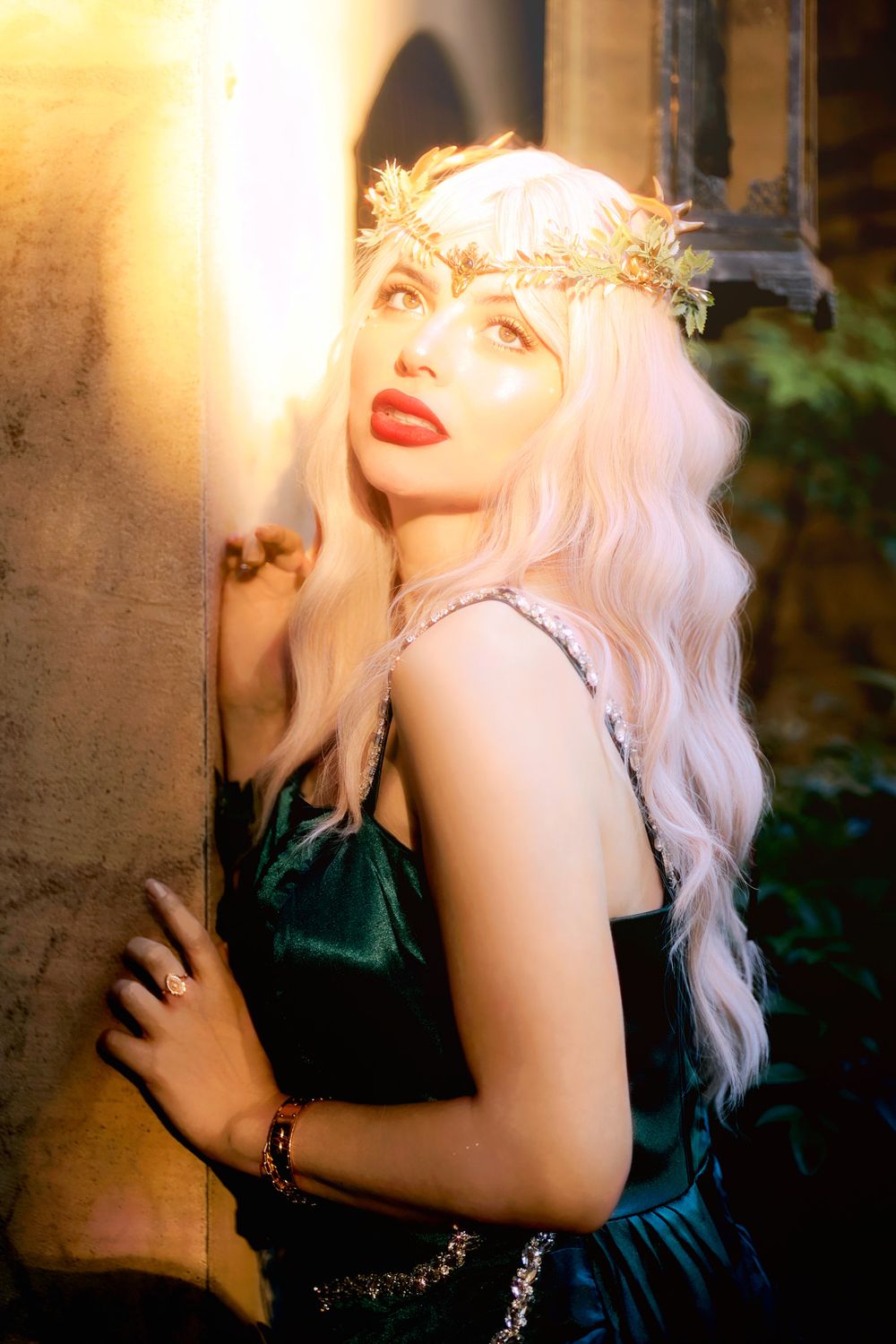 A woman with long white hair and a floral crown poses gracefully in a green dress, bathed in warm golden light.