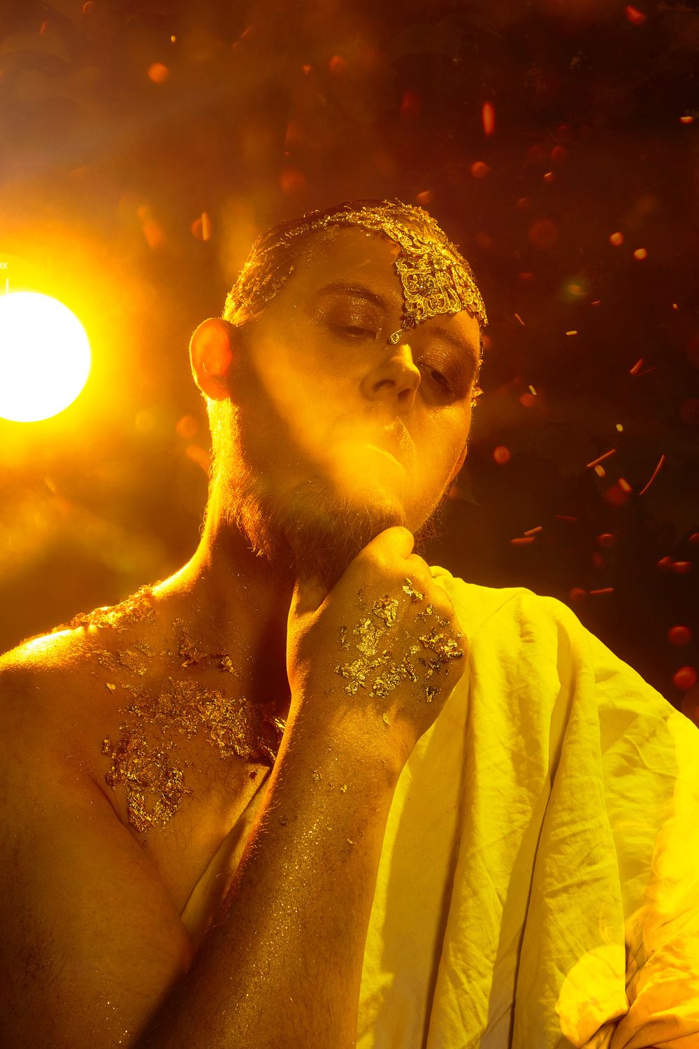 A shirtless man adorned with gold glitter poses dramatically under warm orange stage lighting with confetti falling around him.