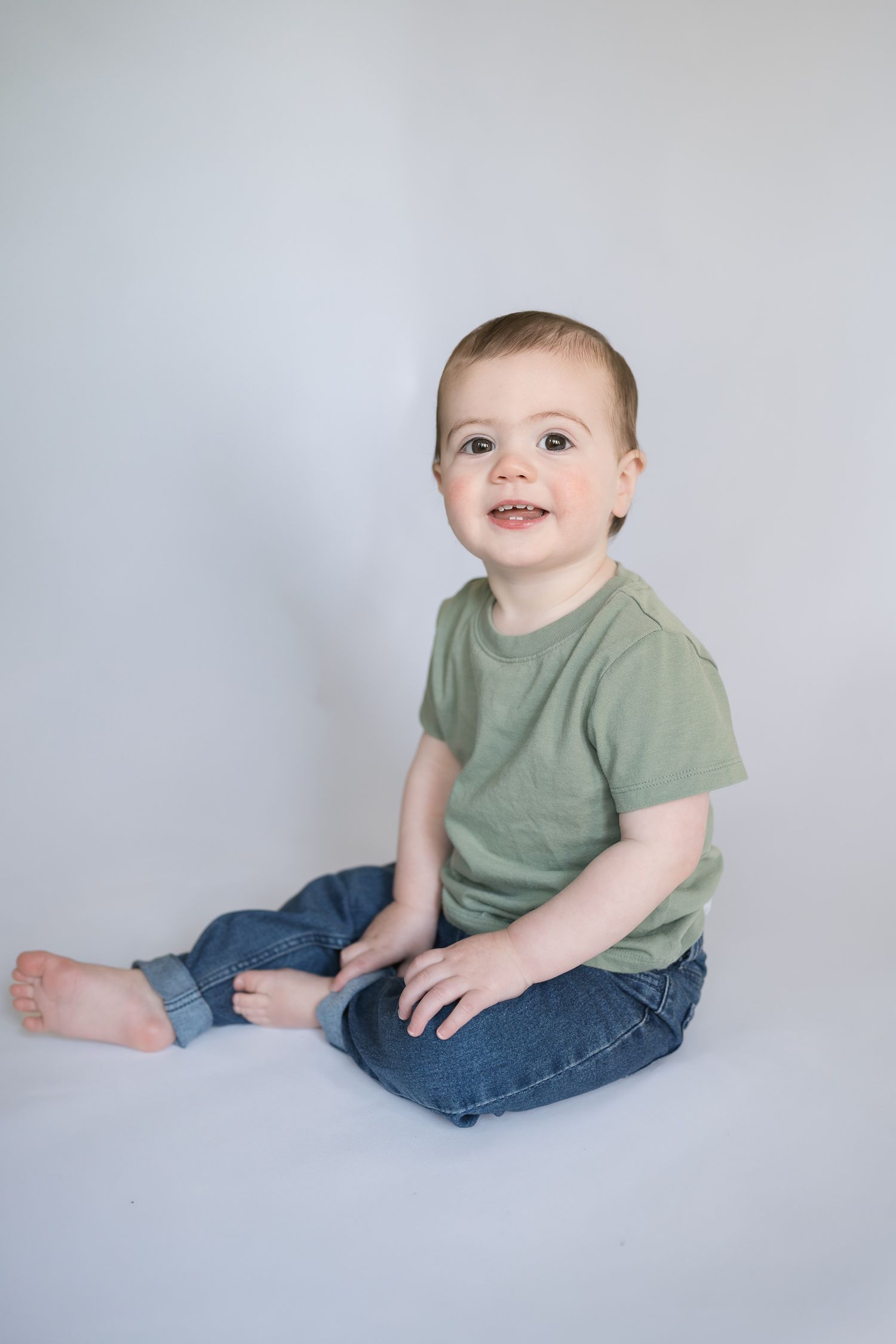 Smiling baby boy in green t-shirt and jeans sitting on white background, adorable infant portrait photo.