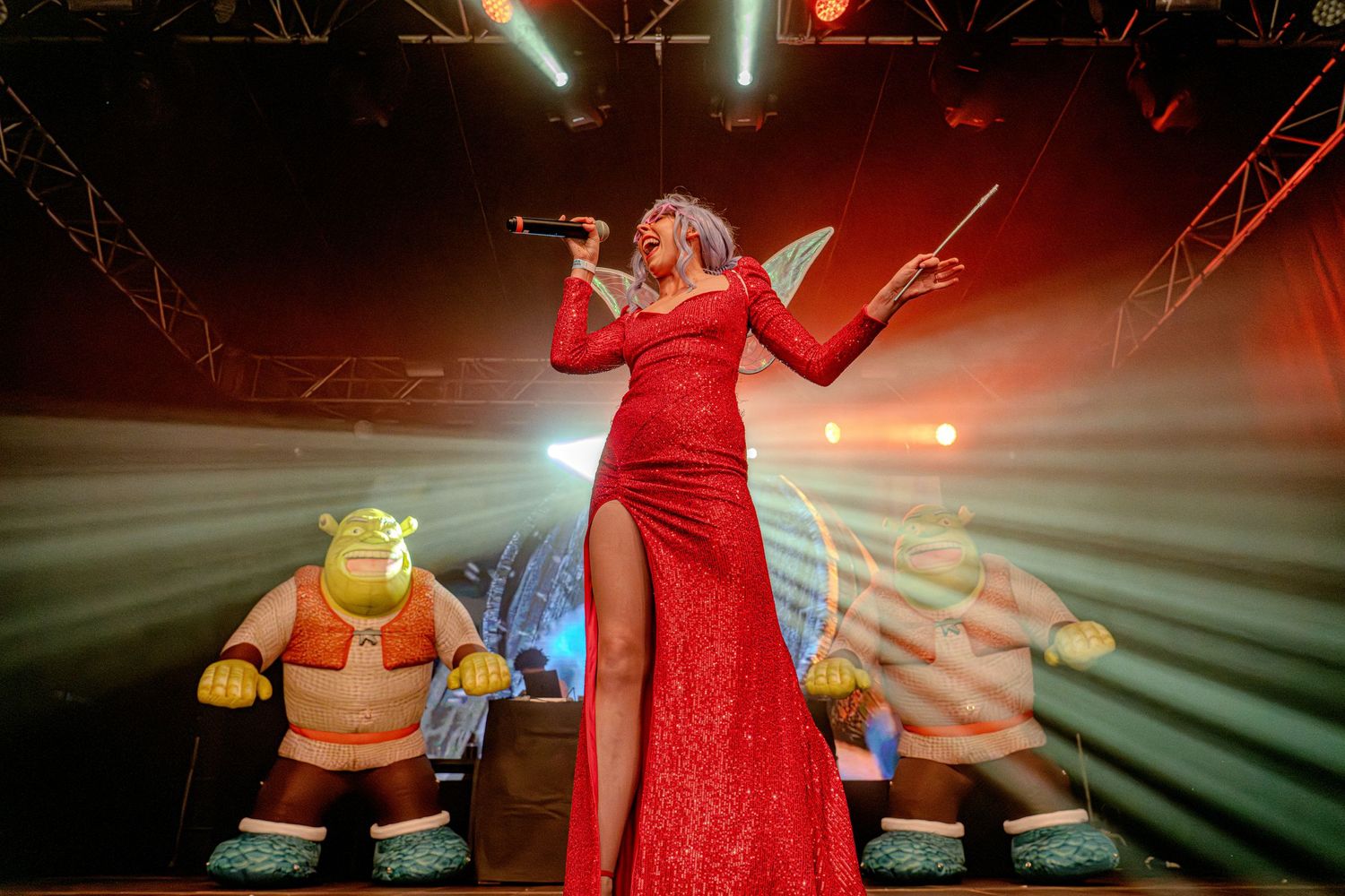 Singer in red gown performing on stage with Shrek-themed character decorations and dramatic lighting effects.