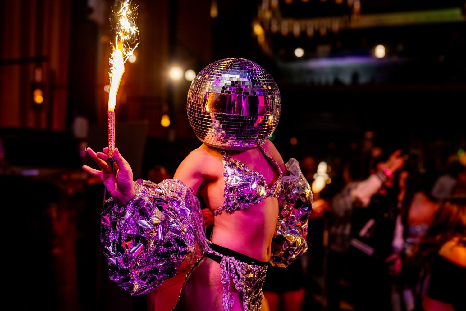 Two disco balls reflect colorful purple lights onto a dark nightclub dance floor with people dancing in the background.