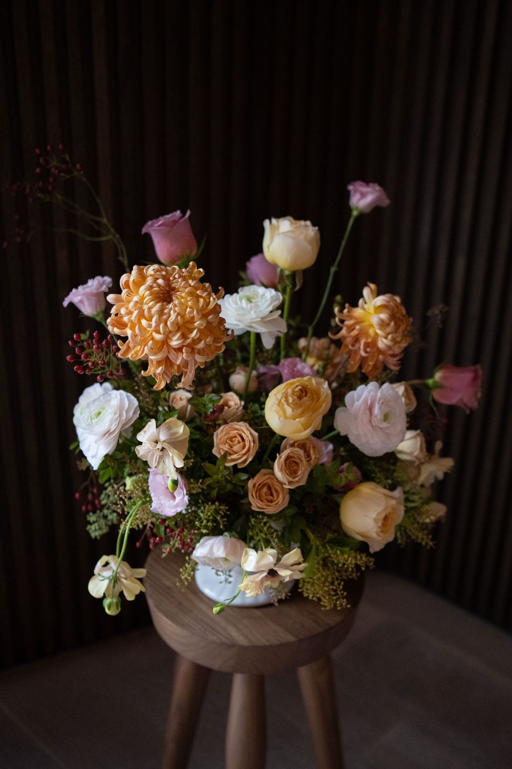 Elegant floral arrangement featuring peach roses and white blooms in a modern brown vessel against a dark background.