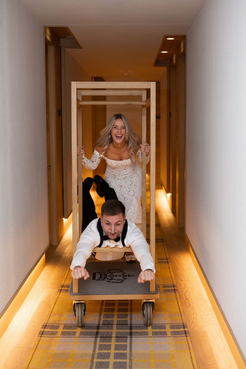 Playful couple crawls through a brightly lit hotel hallway with plaid carpeting and warm wall sconces.