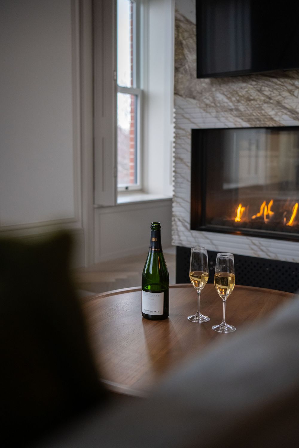 A romantic setting with champagne bottle and two glasses by a lit fireplace near a window. The Royal Hotel Prince Edward County
