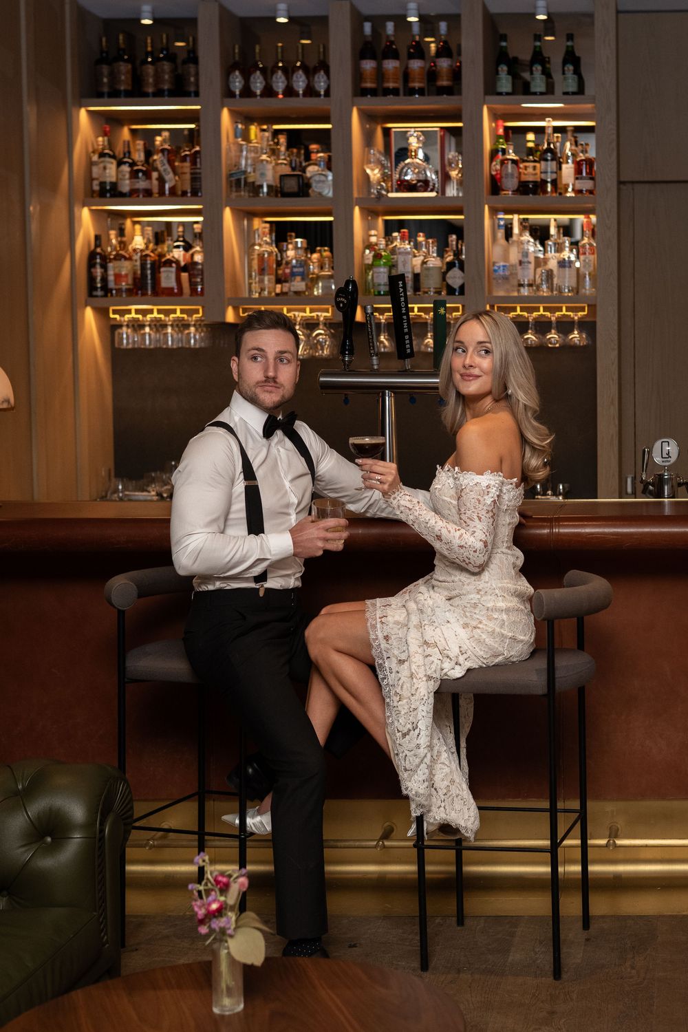 A couple in formal attire poses playfully at a luxurious bar counter with ambient lighting.