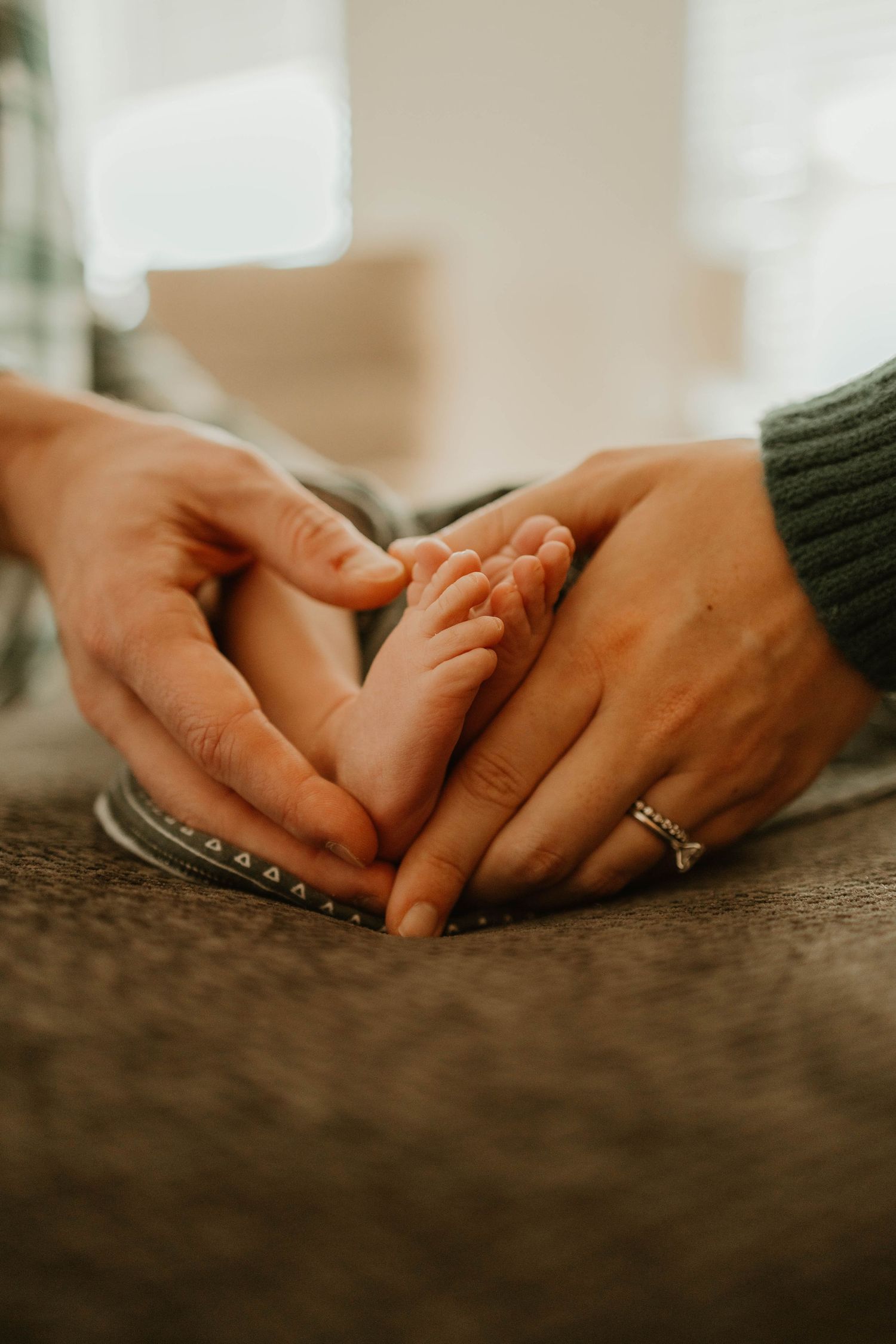Adult hands tenderly holding baby's tiny foot on textured surface, with engagement ring visible.