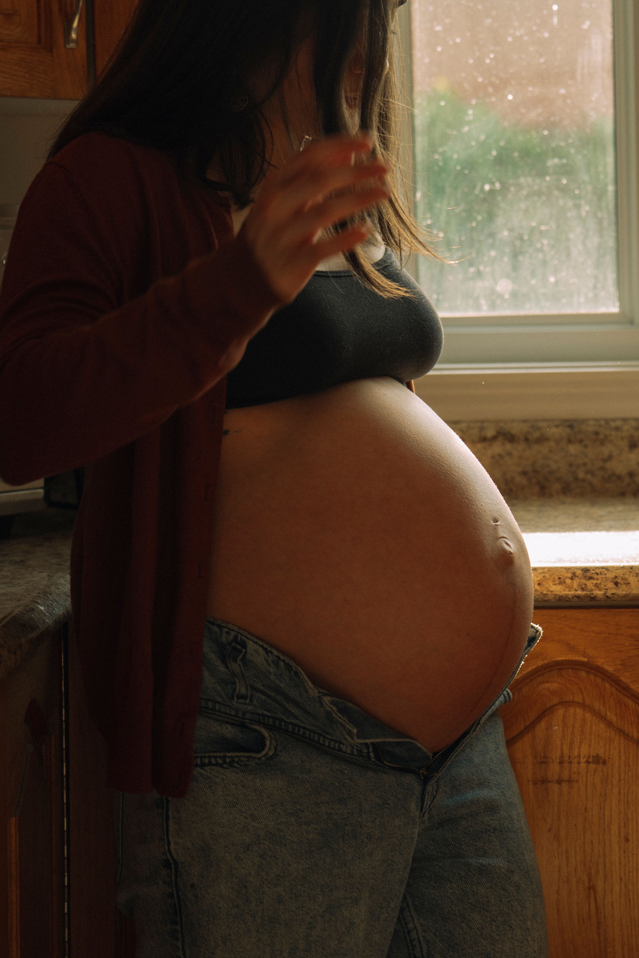 Pregnant woman in kitchen showing baby bump, wearing jeans and red cardigan, standing by window.