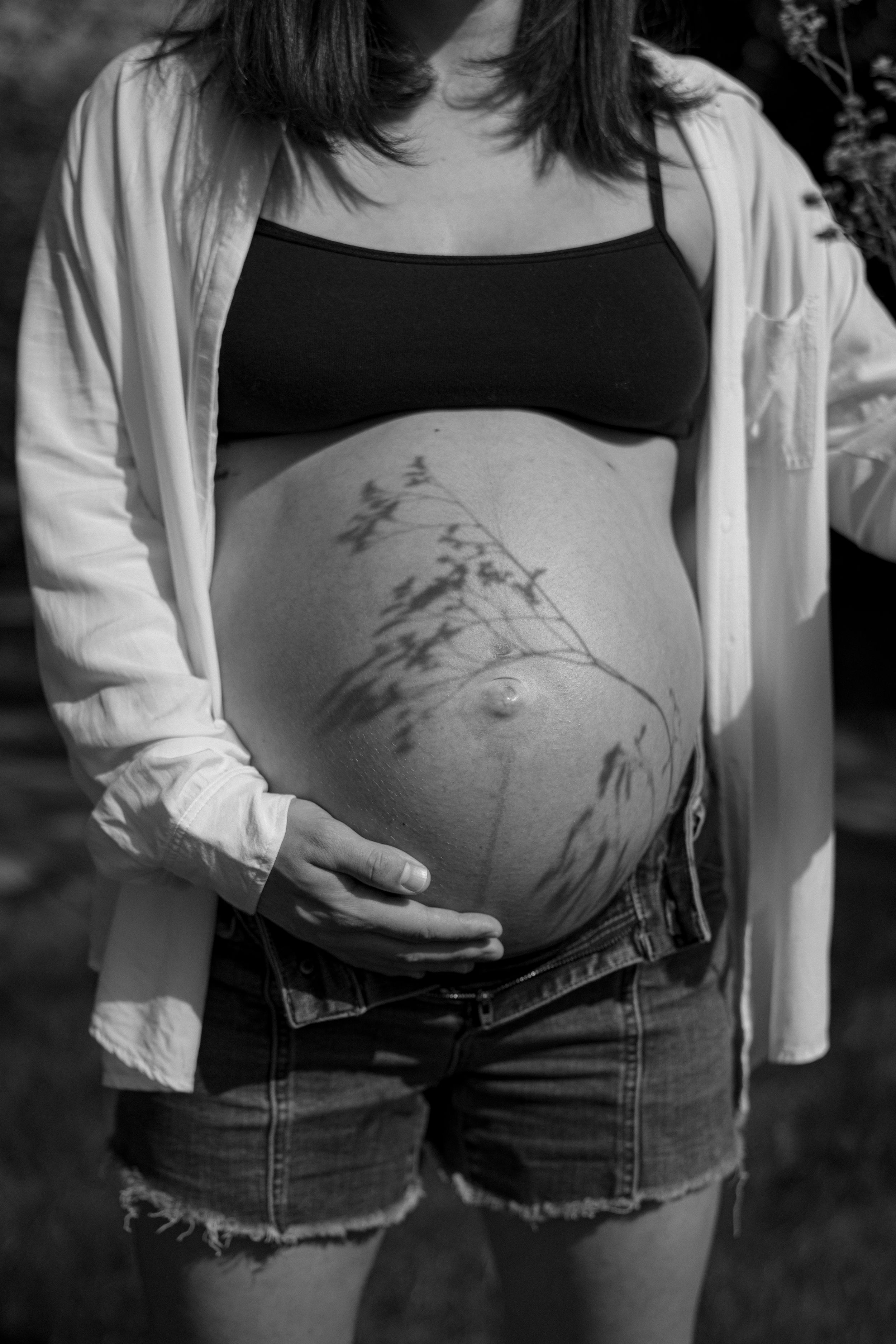 Black and white maternity photo of pregnant woman cradling bare belly with floral tattoo, wearing denim shorts and open white shirt.