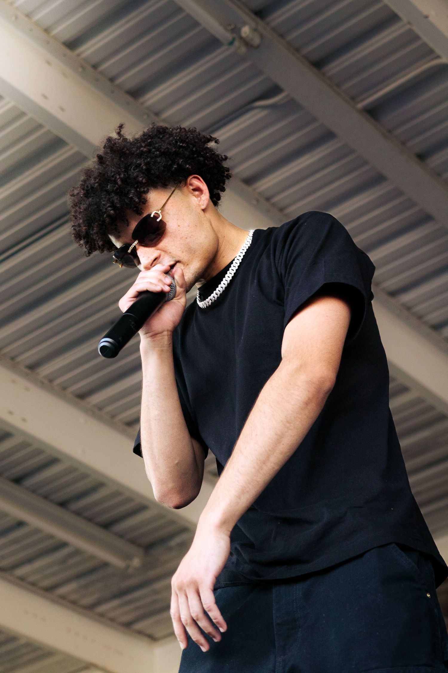 Young male performer singing into microphone wearing black tee and sunglasses.