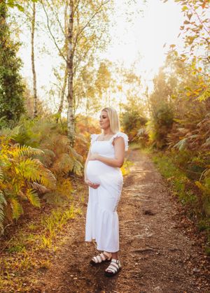 A pregnant woman in a white dress stands on a sunlit forest path surrounded by autumn foliage.