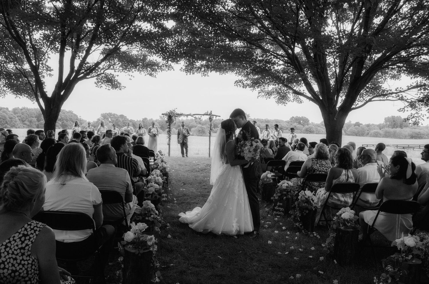 Outdoor wedding ceremony under large trees with guests seated on white chairs watching couple at rustic wooden arch.