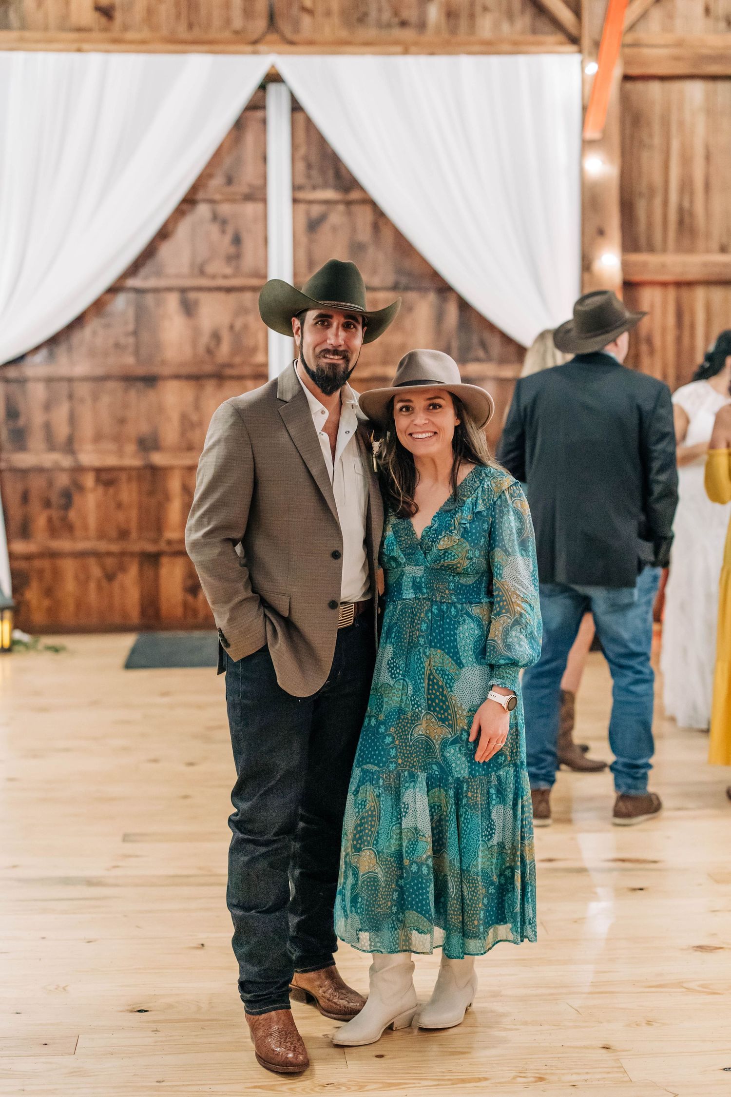 Couple in western attire posing in a rustic barn wedding venue with white draping in the background.