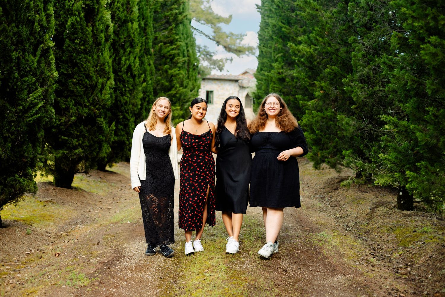Four friends in dark dresses stand together on a tree-lined path.