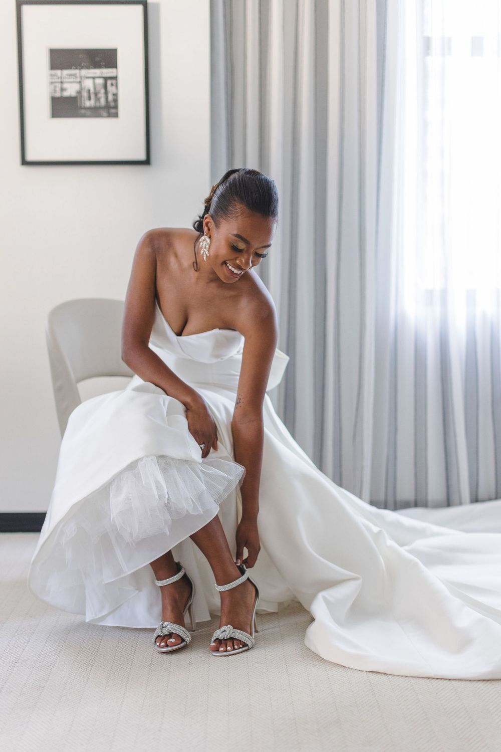 Joyful bride in strapless wedding gown adjusting her shoes in bright modern room.