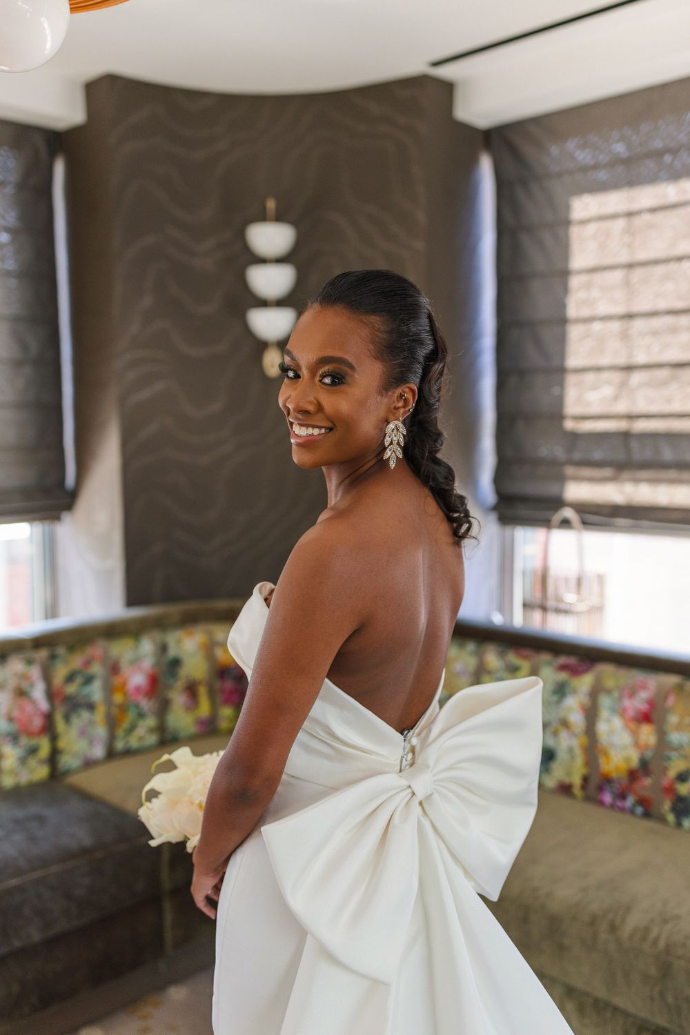 Elegant bride in white gown with dramatic bow detail stands in modern venue setting.