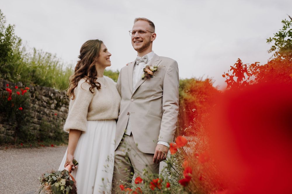 A couple smiles at each other in a garden with red flowers and autumn foliage in the background.