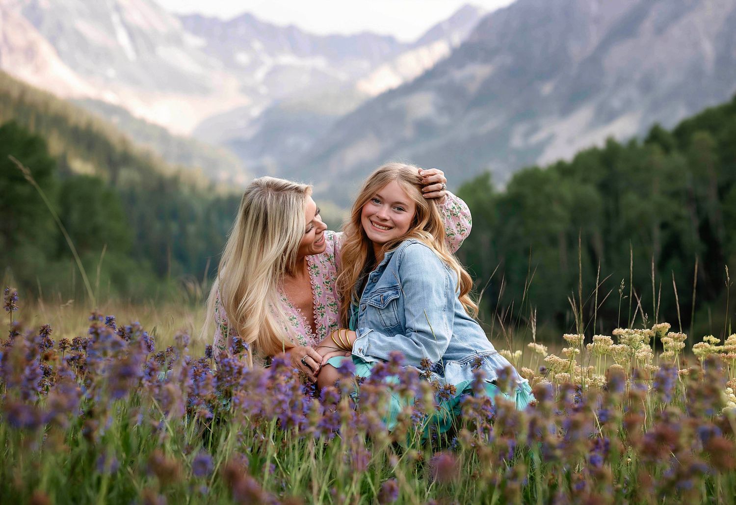 A sweet mother looks at her beautiful daughter and smiles, sitting in the wildflowers- photo taken by Kelly Elizabeth in Aspen CO