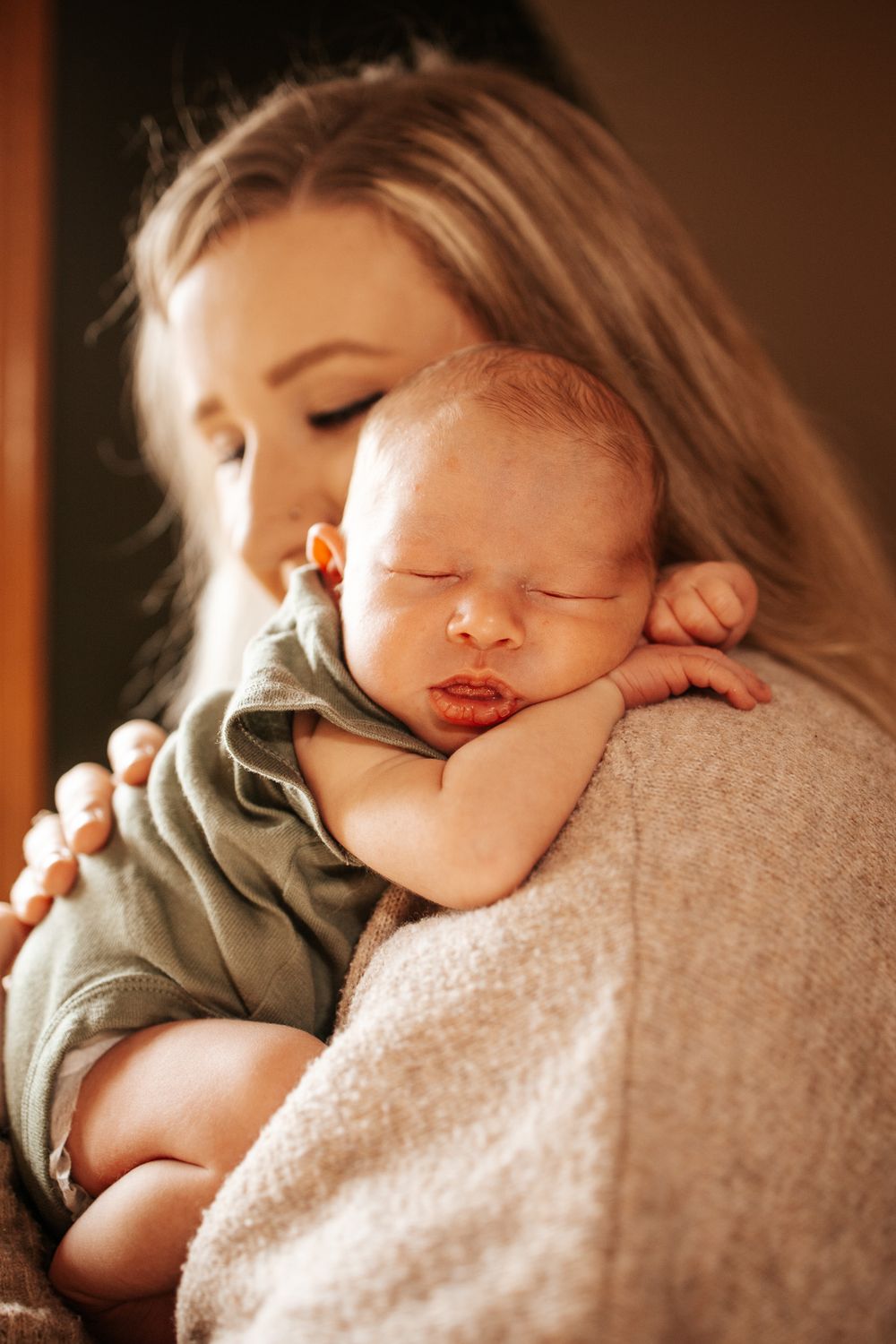Mother tenderly holds sleeping newborn baby on her shoulder in warm natural light.