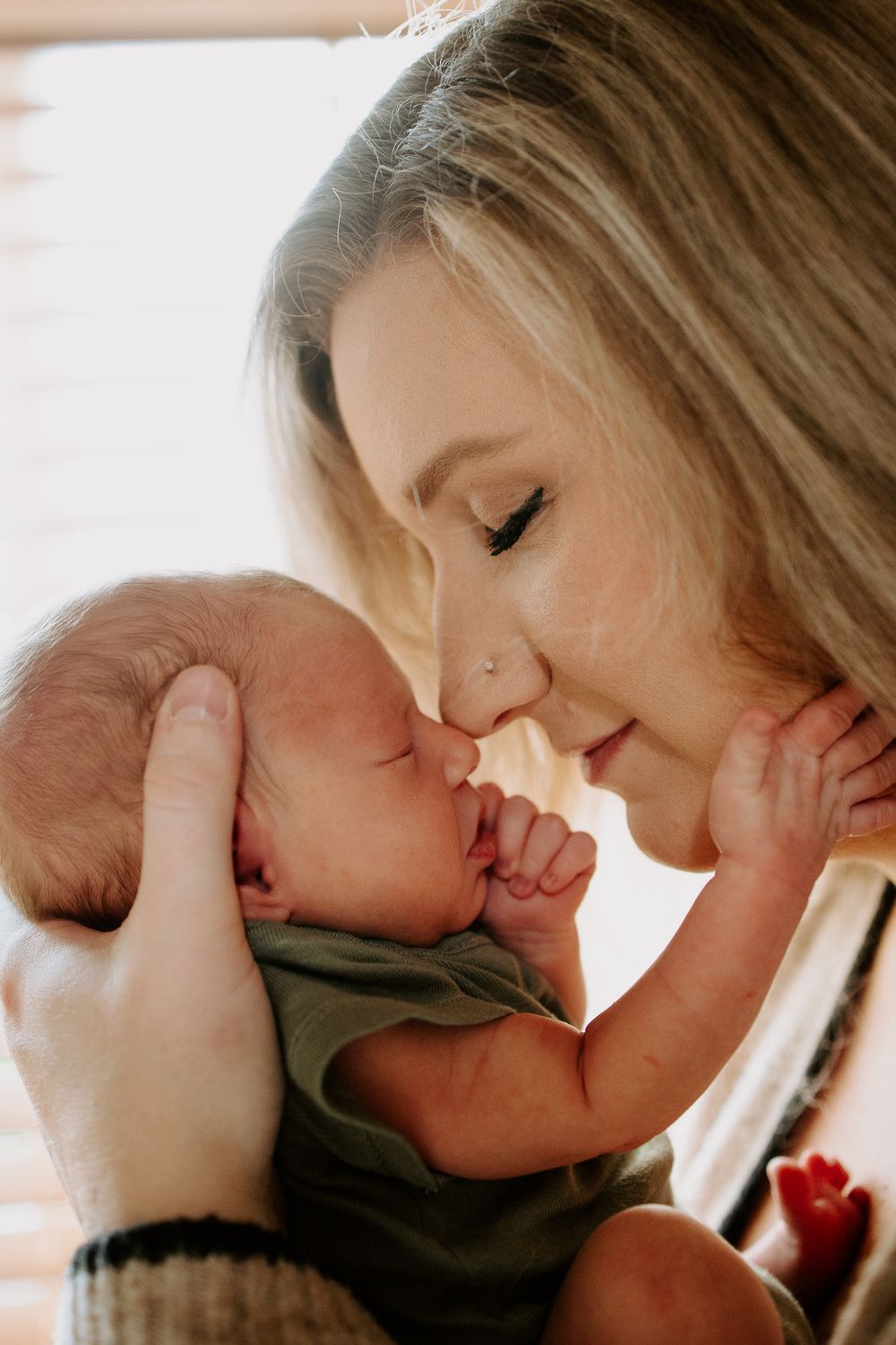 A tender moment as a blonde mother gently touches noses with her sleeping newborn baby, bathed in soft natural light.