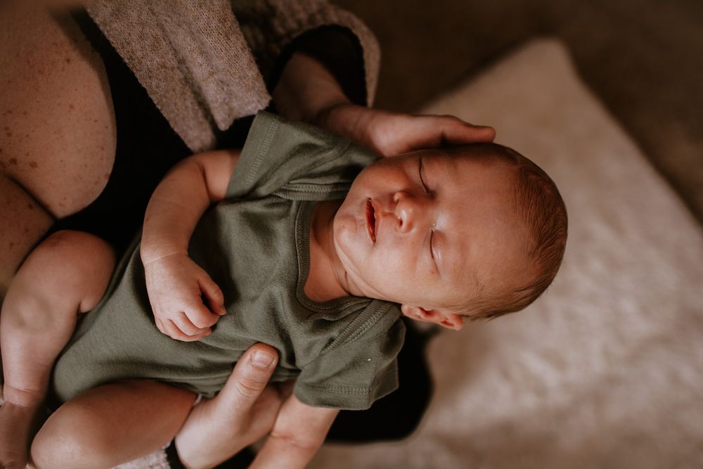 Newborn baby in sage green onesie resting in parent's arms with eyes closed in warm natural light.