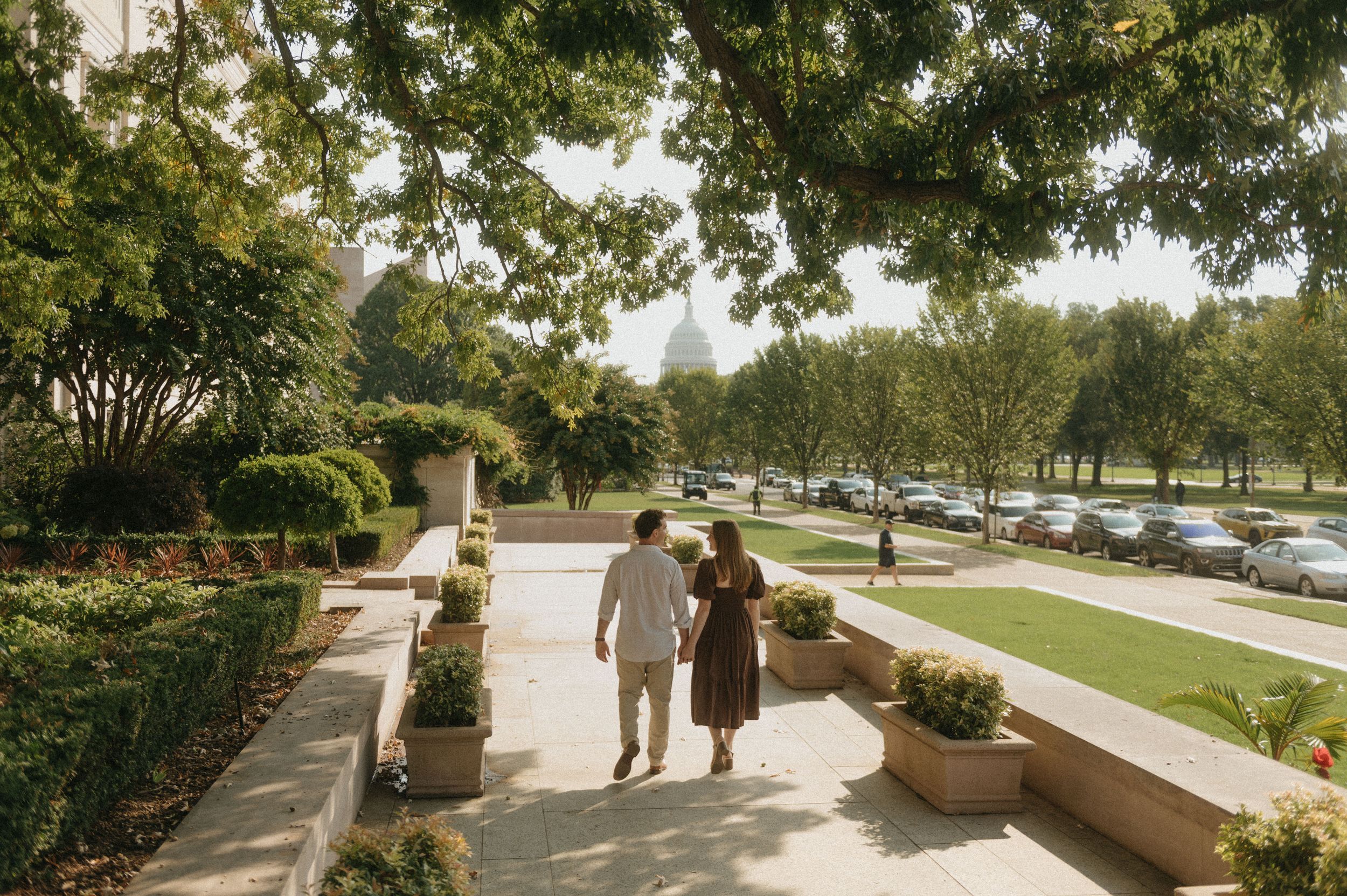 People stroll along a winding pathway through a landscaped garden area with trees and planters on a sunny day.