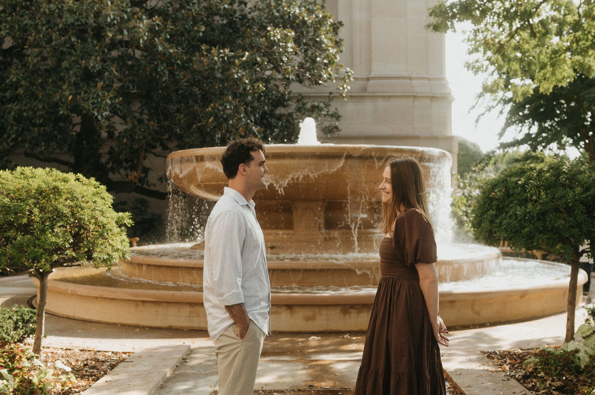 Two people stand facing each other in front of an elegant stone fountain in a sunlit garden setting.