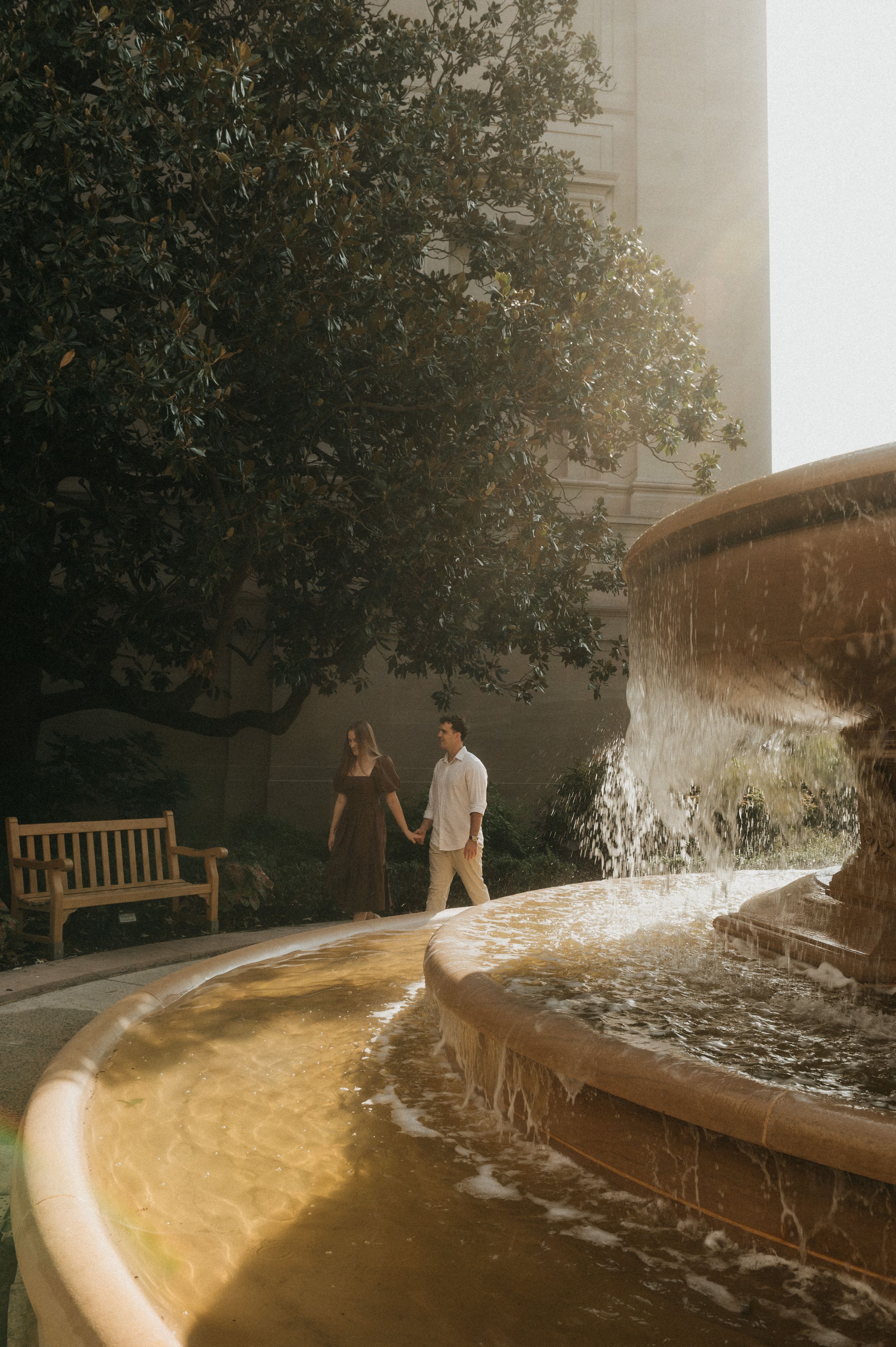 Water fountain creates a dramatic splash effect along a curved stone wall in a garden setting at sunset.