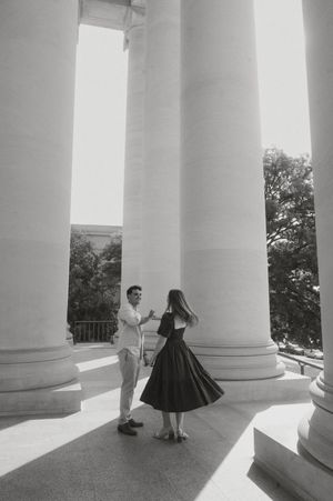 A couple in formal attire dances between white classical columns in an elegant architectural setting.