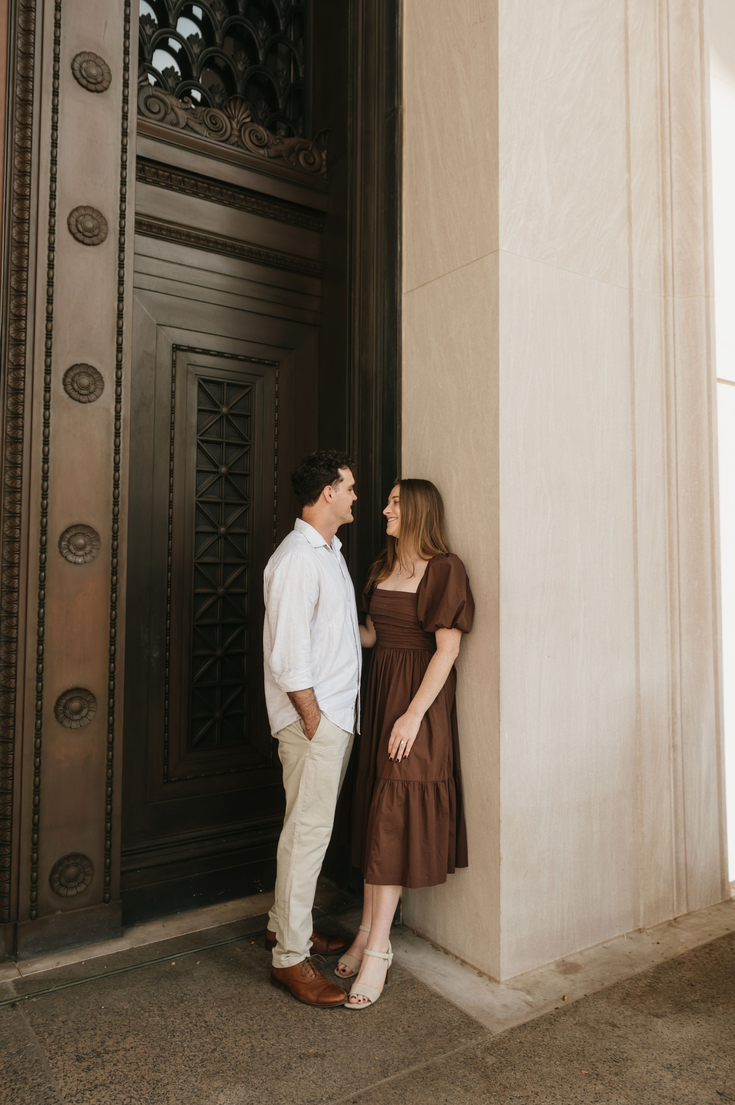 A couple in contrasting brown and white outfits share an intimate moment against an ornate doorway and stone wall.