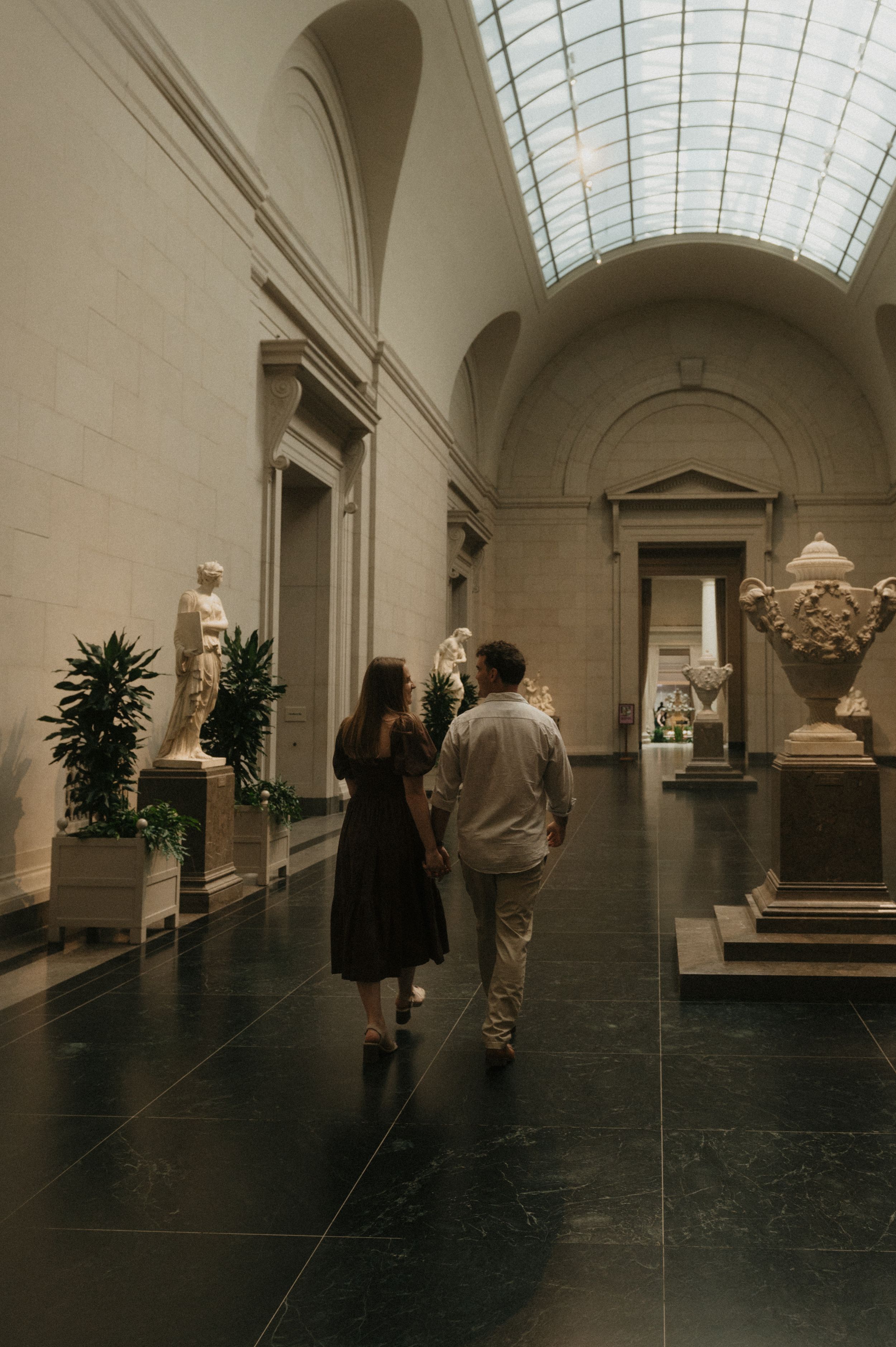 Art gallery visitors explore a long hallway with marble sculptures and skylit ceilings in a classical museum setting.