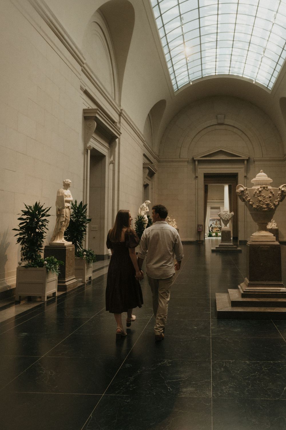 Art gallery visitors explore a long hallway with marble sculptures and skylit ceilings in a classical museum setting.