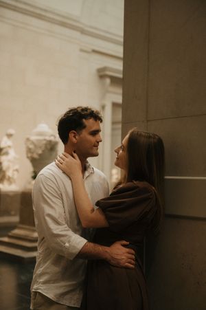 A romantic couple embraces intimately in a dimly lit museum or gallery space with white walls and sculptures.