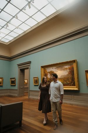 Two people walk through an art gallery with turquoise walls and wooden floors displaying landscape paintings.