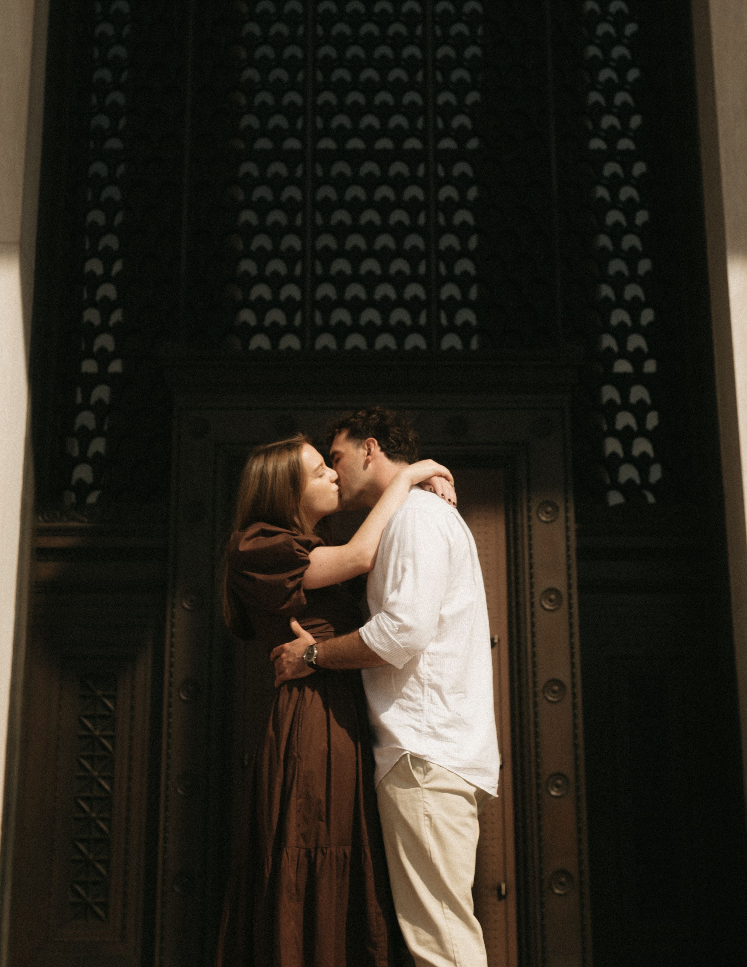 A romantic couple embraces in front of an ornate dark wooden door with decorative patterns.