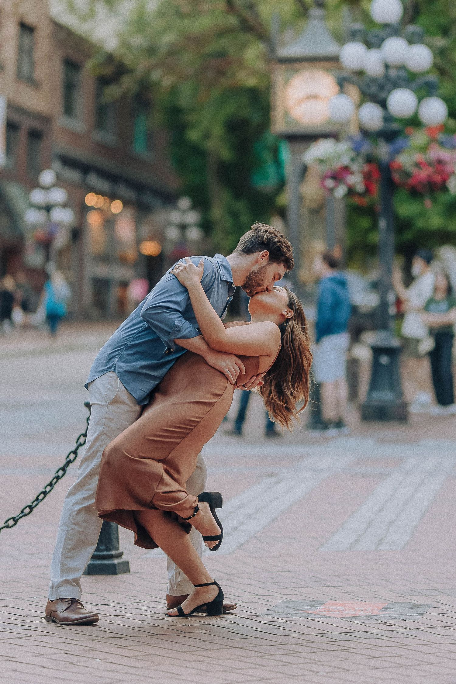 A couple shares a romantic dip-style dance pose on a brick sidewalk with decorative lampposts and flowers in the background.