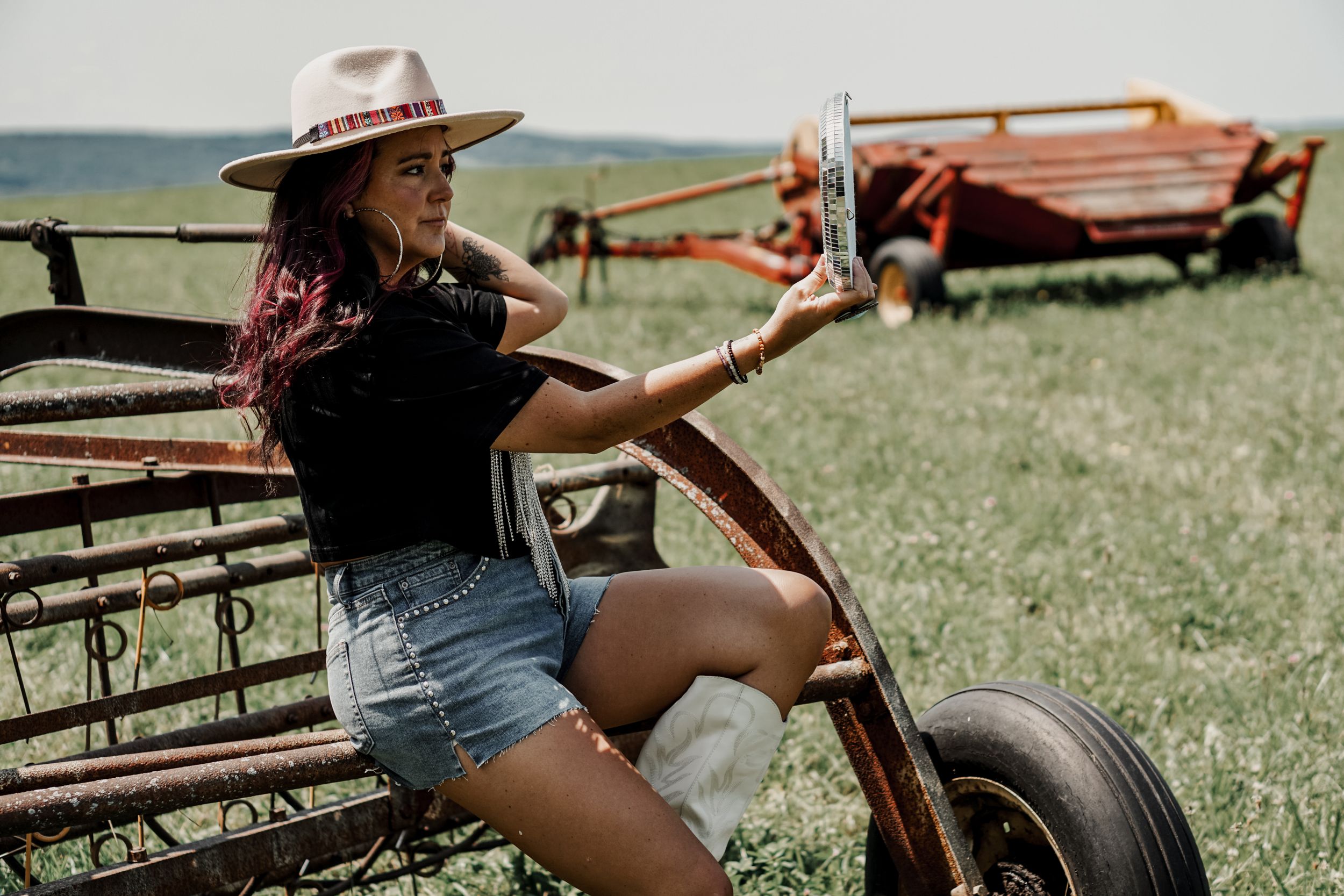 Casual summer outfit with denim shorts and hat against rustic farm equipment backdrop.