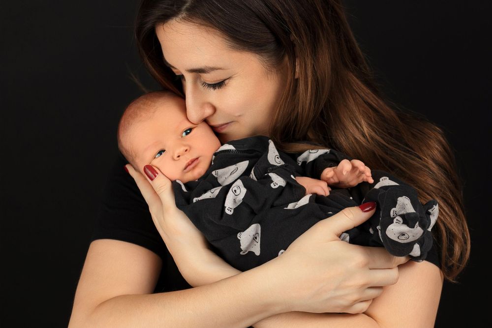 Mother in black patterned top embracing newborn baby against dark background.