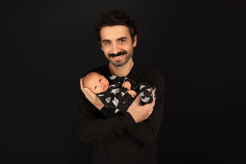 Father in black shirt holding newborn baby wearing patterned onesie.
