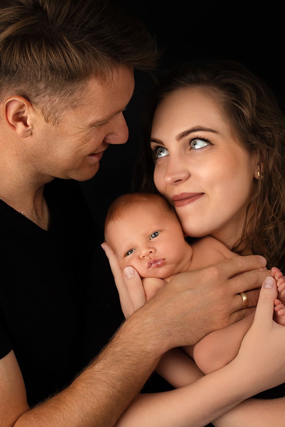 Intimate family portrait against black background featuring newborn baby nestled between parents.