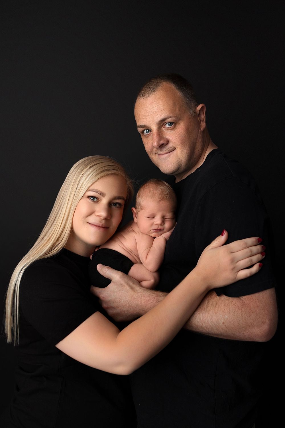 Studio portrait of a family in black clothing on dark background with their sleeping newborn baby.