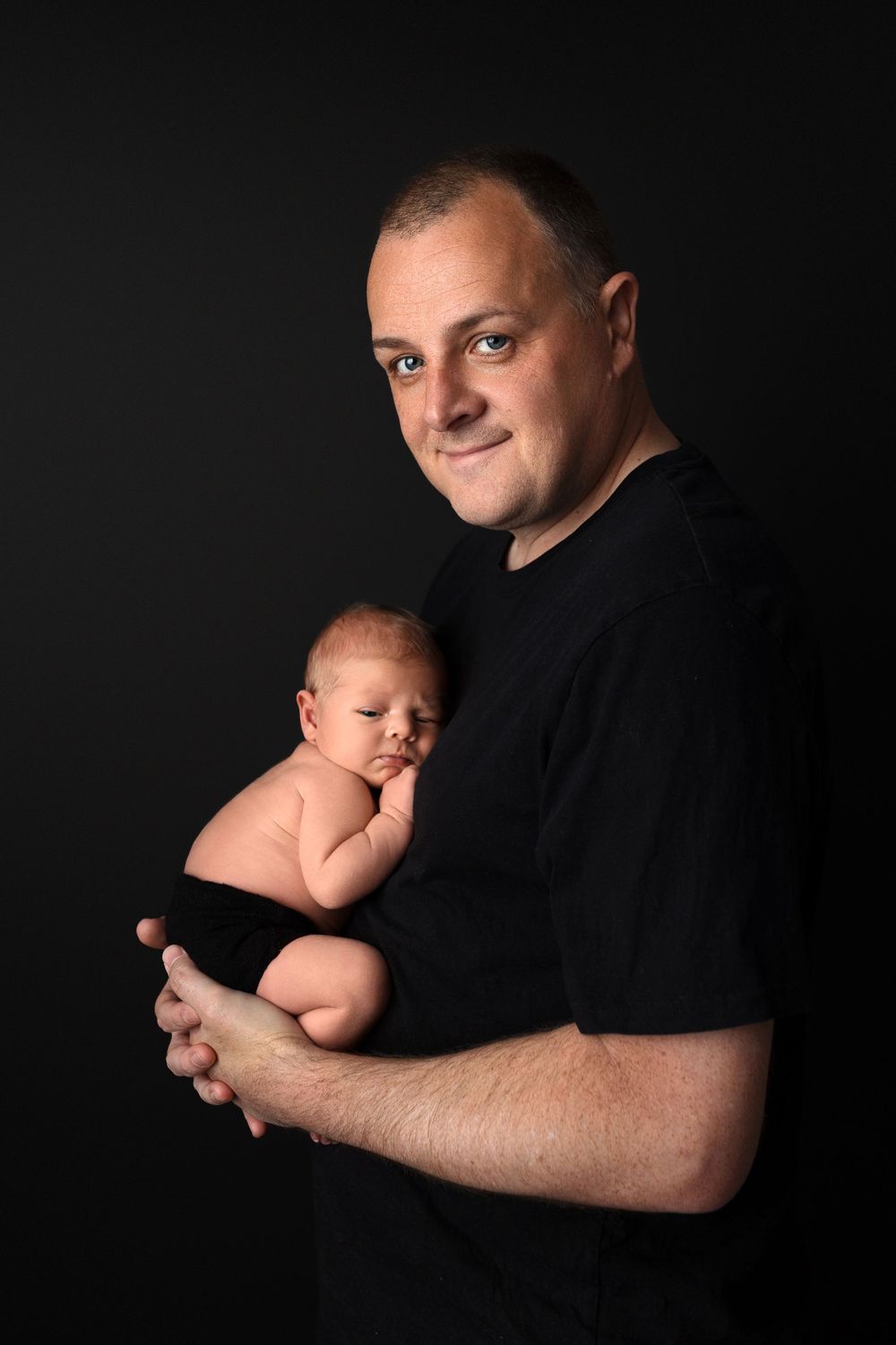Studio portrait of someone in black shirt tenderly holding a sleeping newborn baby against dark background.