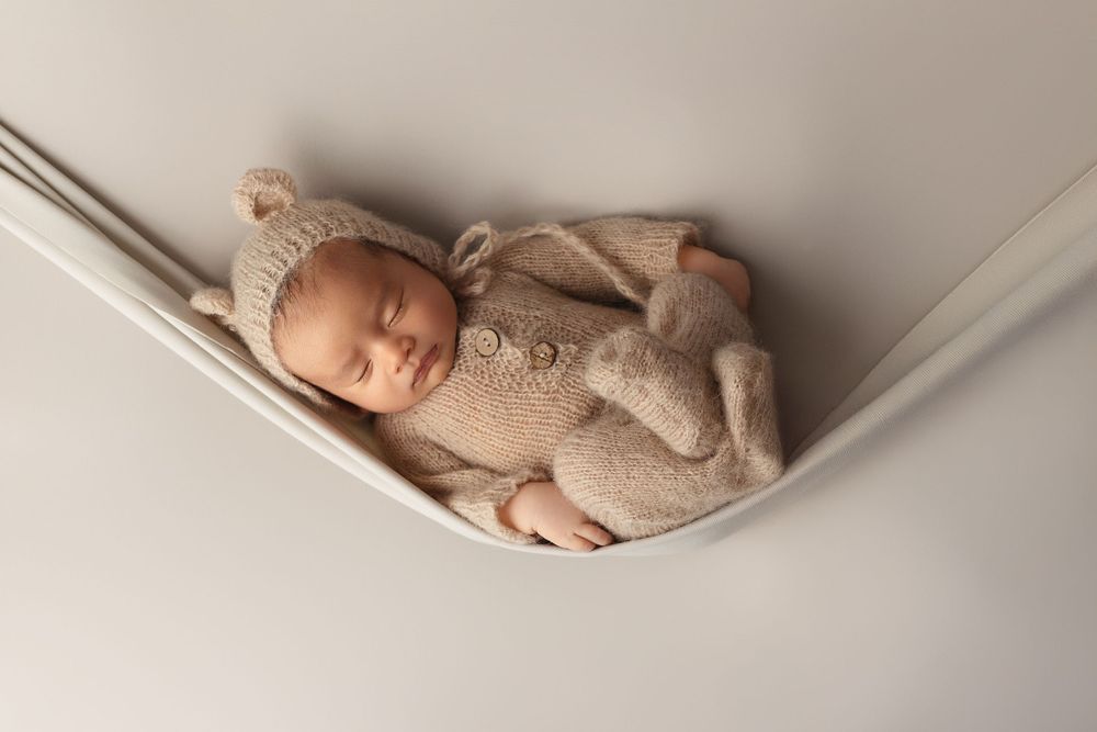 Peaceful newborn in beige knit outfit sleeping in suspended fabric hammock.