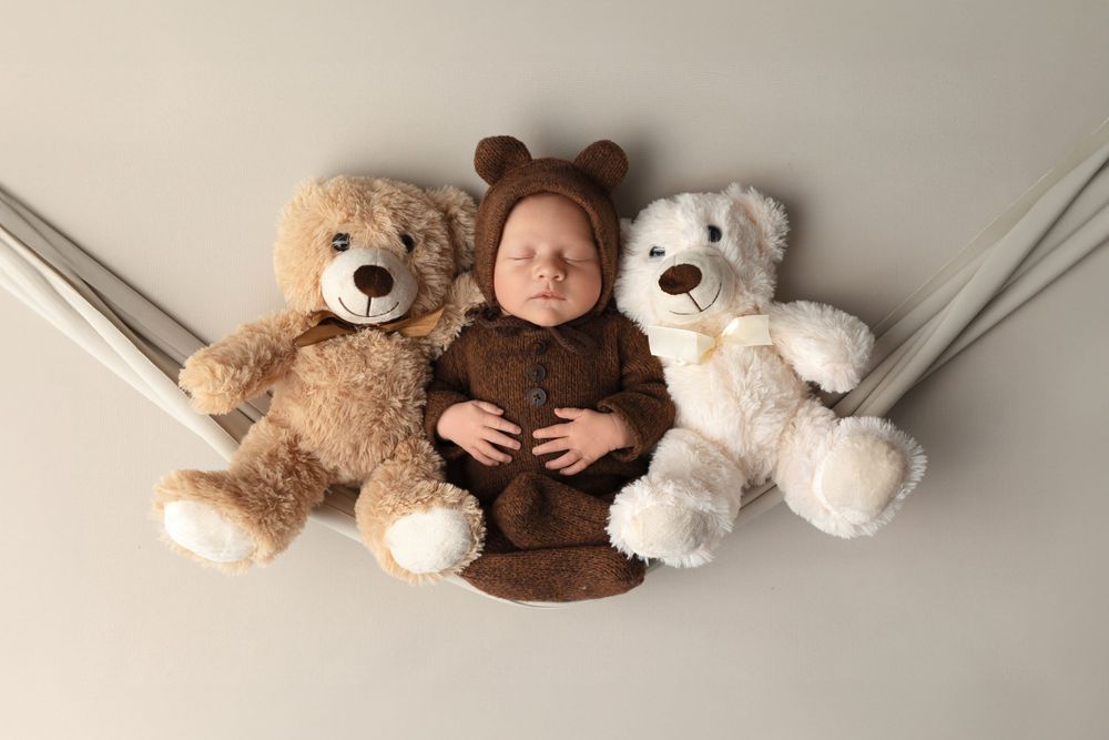 Newborn in brown bear outfit lying between two plush teddy bears on gray background.