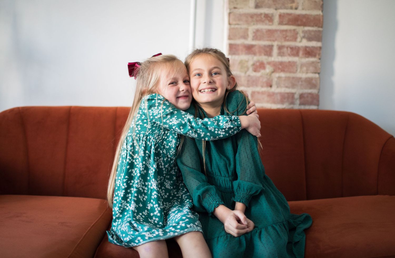 Two sisters in green dresses hugging and smiling on a red sofa indoors.