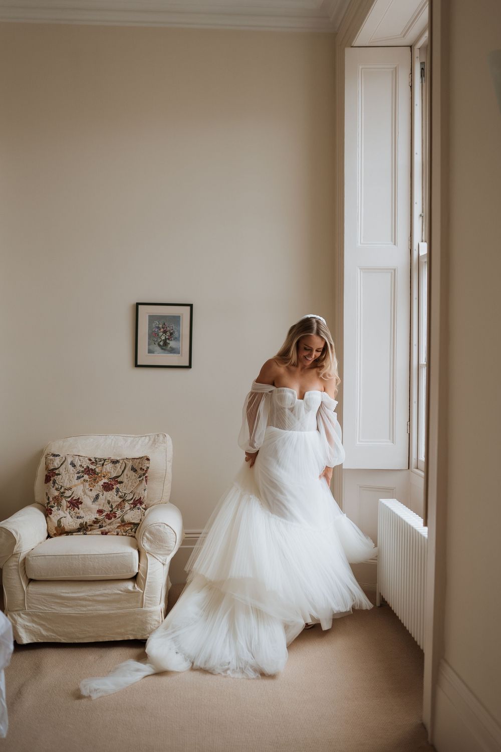 Elegant off-shoulder wedding dress with layered tulle skirt shown in light-filled room with vintage armchair.