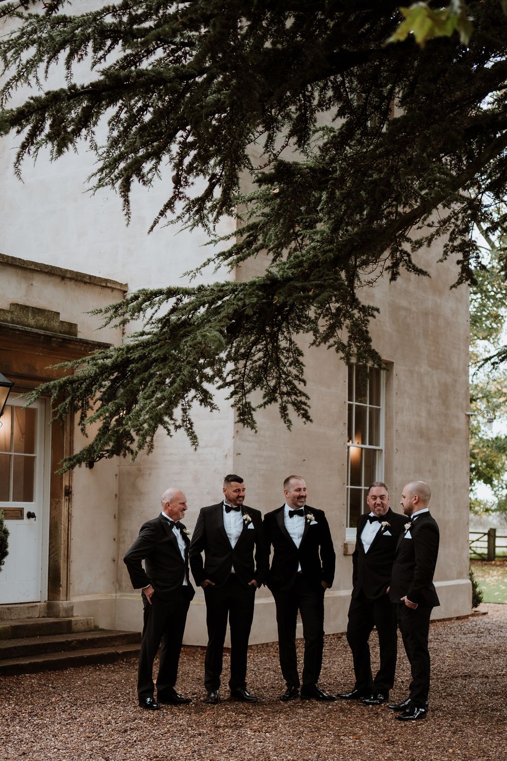 A group of men in black tuxedos stand together smiling outside a white building with evergreen branches overhead.