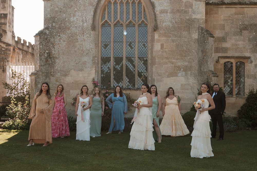 Wedding party poses together outside historic stone church with gothic windows in golden evening light.
