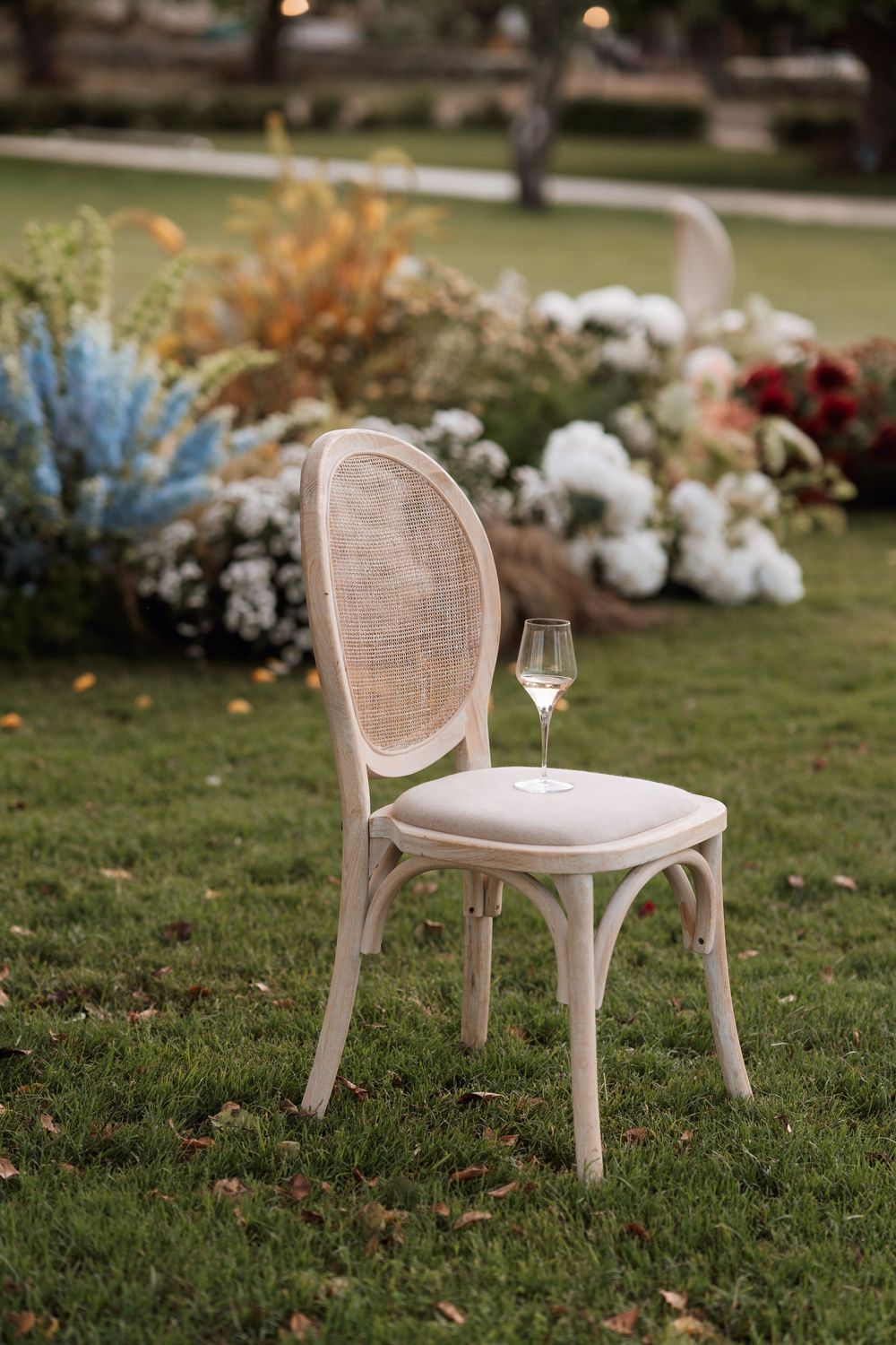 Vintage cane chair with plush cushion sits alone on green lawn with floral arrangements in outdoor wedding setting.