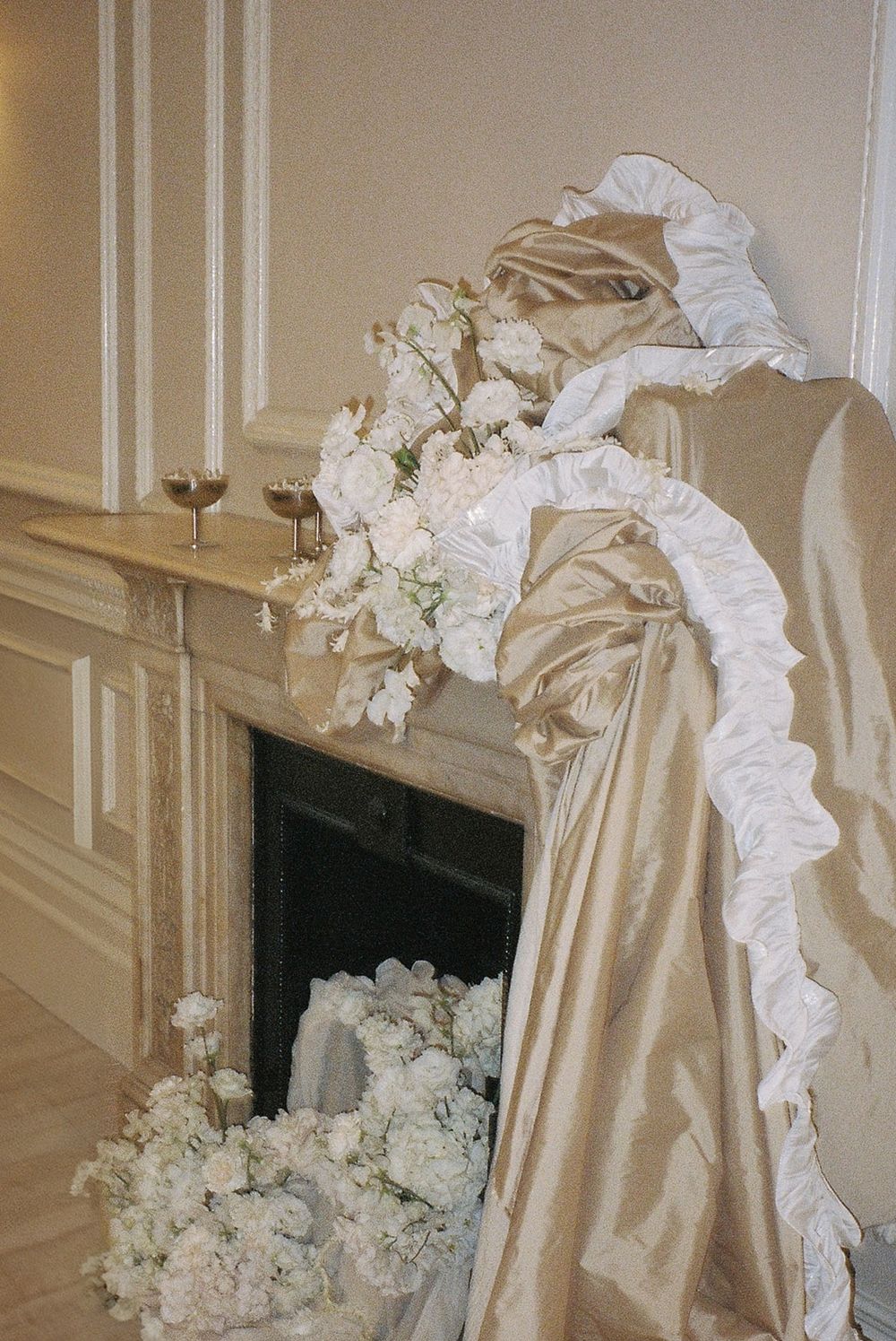 Elegant cream colored drapery with white ruffled trim and floral arrangements adorning a classical fireplace mantel.